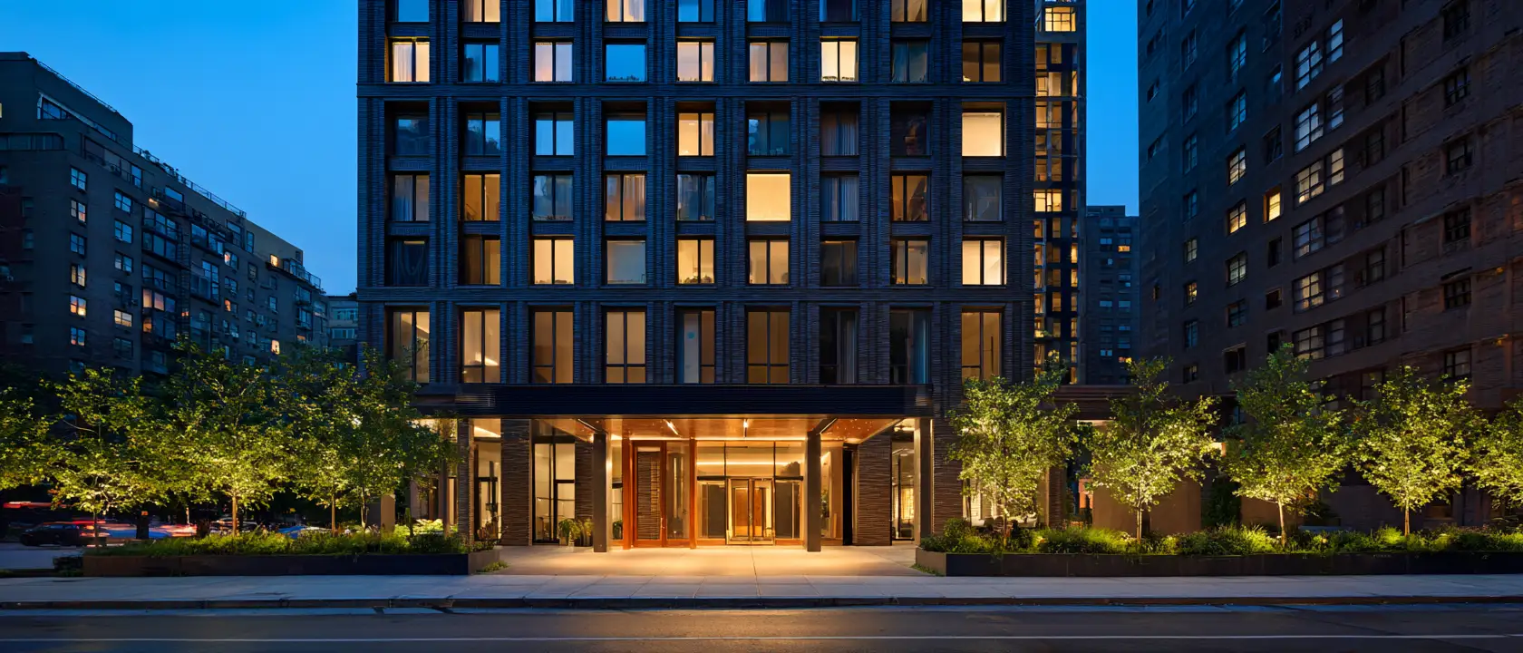 A modern residential building at dusk, its lit entrance and windows warm against a deep blue sky